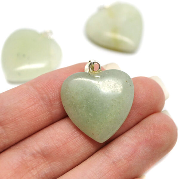 A hand holding a small light green jade carved heart pendant against a white background